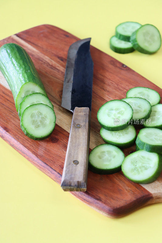 黄瓜(Cucumis sativus)和菜刀在木制砧板上的图像，整个水果和切片显示绿色的皮，白色的肉和白色的种子，黄色背景，高架视图，重点在前景